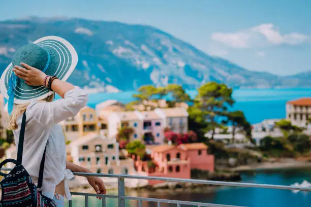 Tourist woman wearing blue sunhat and white clothes enjoying view of colorful tranquil village Assos on sunny day. Stylish female visiting Kefalonia in summer time on Greece travel vacation.