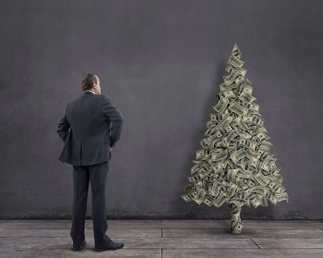 Rear view of businessman standing on wooden floor with flowing one hundred-dollar bills in the shape of Christmas tree.