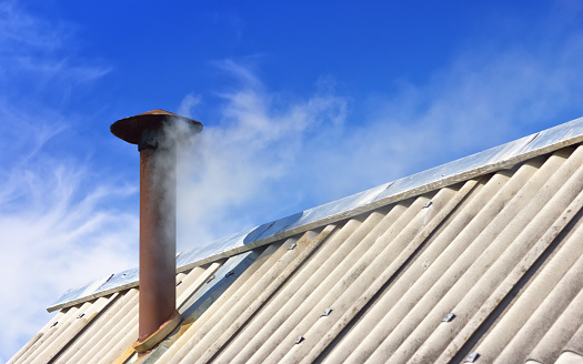 Rusty chimney pipe on a slate covered roof of an old house. White Smoke from the chimney against the blue sky with clouds. Abstract concept background with space for copy, selective focus.