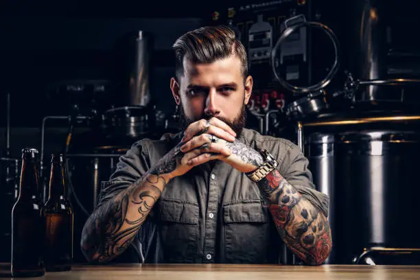 Photo of Portrait of a pensive tattooed hipster male with stylish beard and hair in the shirt in indie brewery.