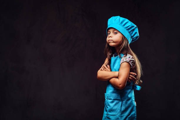 不機嫌そうな少女を着た青クック制服立っている組んだ腕を持つ。ダーク テクスチャ背景上に分離。 - pan saucepan kitchen utensil isolated ストックフォトと画像