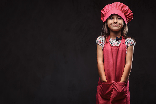 Cute little girl dressed in pink cook uniform holds arms in pockets. Isolated on a dark textured background.