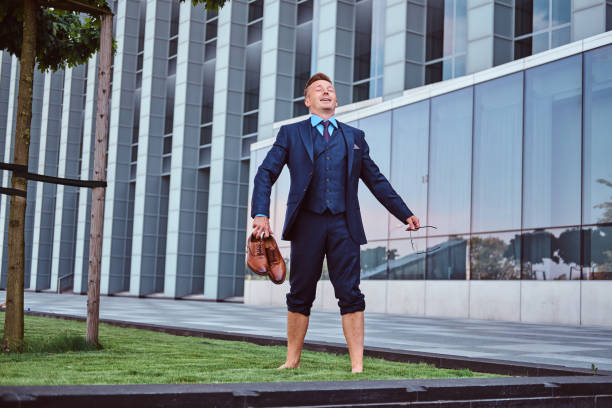 istock Confident businessman dressed in elegant suit holds his shoes enjoying while standing barefoot on a green lawn against cityscape background. 1071807892