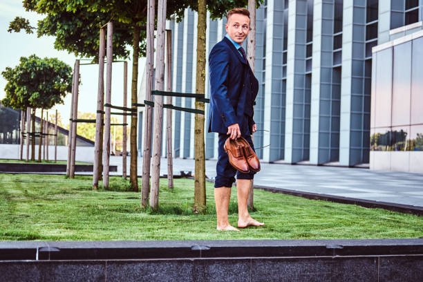 istock Confident businessman dressed in elegant suit holds his shoes while standing barefoot on a green lawn against cityscape background. 1071807926