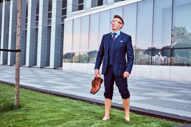 istock Confident businessman dressed in elegant suit holds his shoes enjoying while standing barefoot on a green lawn against cityscape background. 1071808770