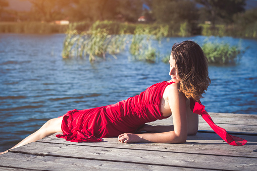 Young brunette caucasian woman wearing elegant red dress is lying on a wooden jetty on a lake on a sunny summer afternoon.