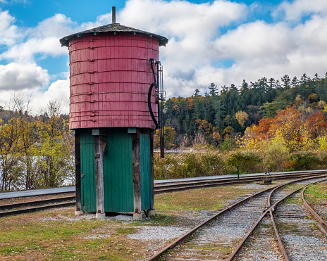 In Wakefield, Quebec, Canada, an old train station water tower stands between two sets of train tracks with autumn trees , blue sky and white fluffy clouds in the background.