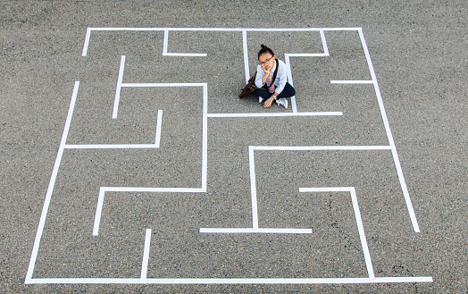 A young business girl and businesswoman wearing business attire and holding a briefcase sits at a dead end in a maze while looking at the camera. She us using her business intelligence and strategy to navigate her new business through the maze, but is struggling at the complexity. She is finding many problems to surmount for his small business to succeed.