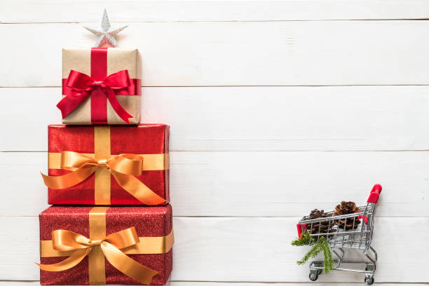 istock Gift box present stack in Christmas tree shape with red-gold ribbon, star, spruce twig, pine cone and shopping trolley on white wood for Xmas boxing day winter holiday seasonal celebration background 1071915402