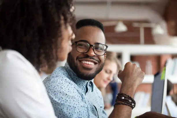 Photo of Smiling African American male employee look at colleague chatting