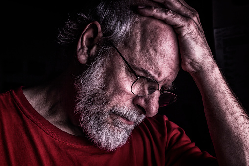 A distraught senior adult man is holding his head with his hand as he looks down.