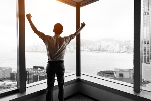 Businessman standing by the window with arms raised