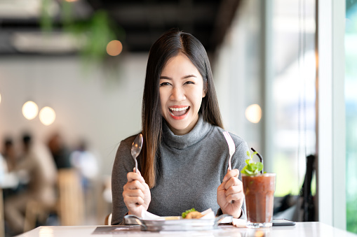 Young attractive asian woman holding fork and spoon feeling hungry, excited, happy and ready to eat healthy food looking at camera in cafe coffee shop. Charming blogger or social influencer concept.