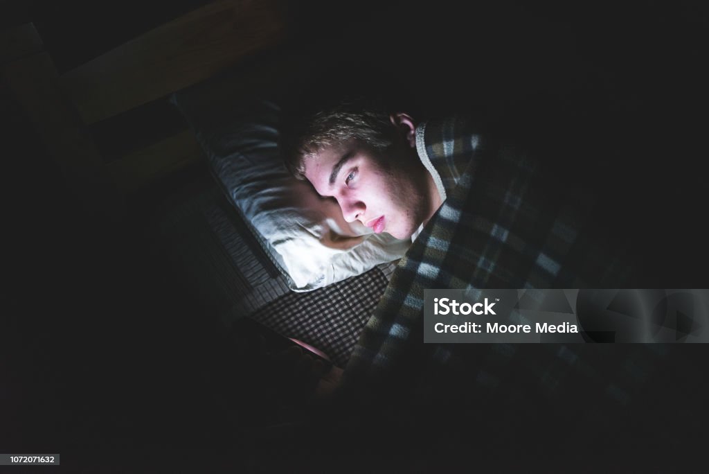 Depressed teenager on his phone in the dark. The image displays a depressed teenager browsing the internet on his mobile phone. He is lying on his bed in the dark. Teenager Stock Photo