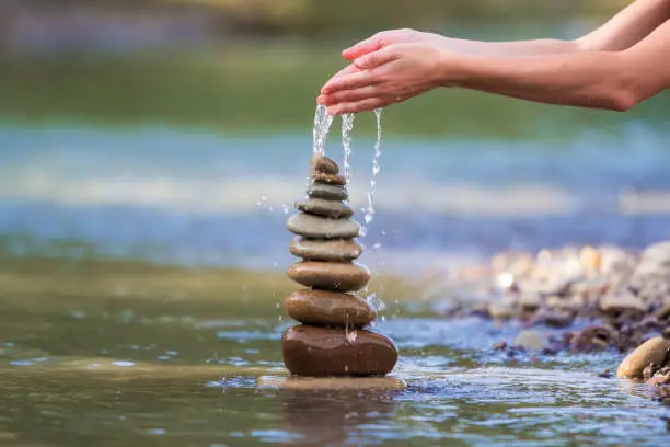 Photo of Close-up abstract image of woman hand pouring water on rough natural brown uneven different sizes and form stones balanced like pyramid pile landmark on blurred blue-green copy space background.
