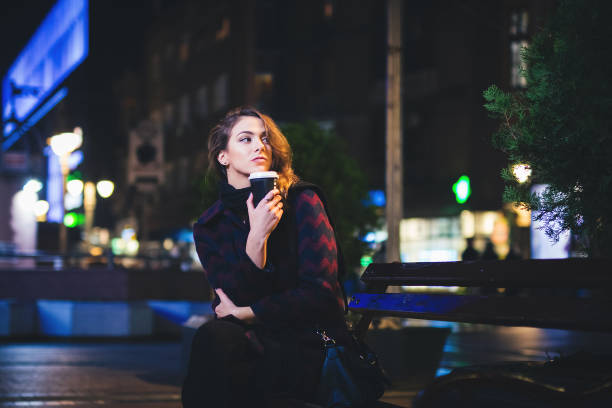 Young beautiful woman sitting on a park bench downtown at night with a coffee to go.
