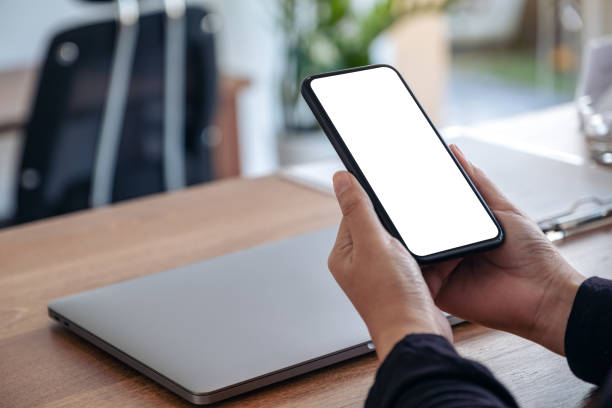 istock Mockup image of hands holding black mobile phone with blank white screen with laptop on wooden table 1072123512