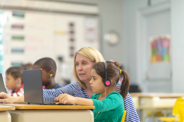 Helping a student A teacher crouches beside the desk of one of her students. The student is working on a laptop in class. They are both looking at the screen as the teacher explains the lesson. teacher classroom child education stock pictures, royalty-free photos & images