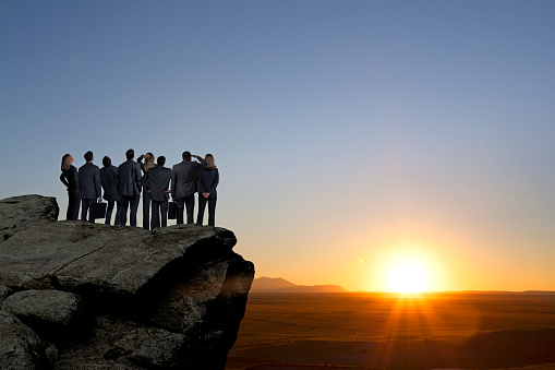 A group of businessman and a businesswoman stand side by side on top of a rocky promontory and look out over a vast plain as the sun is about to set on the horizon in the distance.  She places her hands behind her back as he carries his briefcase.