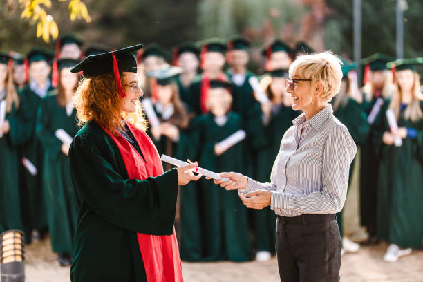 Happy senior professor giving diploma to female student on graduation day. Happy female student talking to her professor while receiving certificate on graduation day. alumni stock pictures, royalty-free photos & images