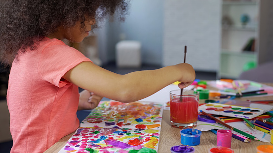 Cute curly African American girl putting paintbrush into water, leisure time