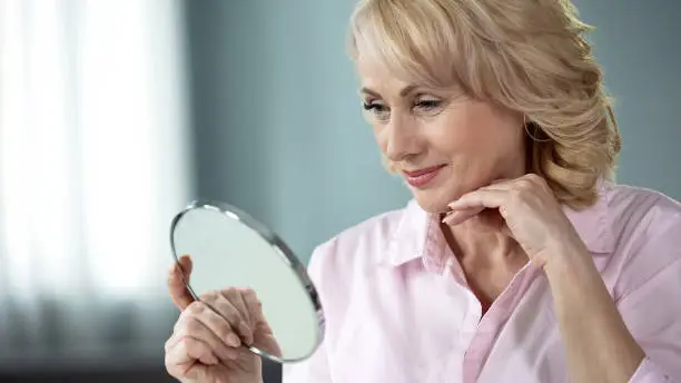 Lady over 50 looking at herself in mirror, rejoicing at result of skin lifting