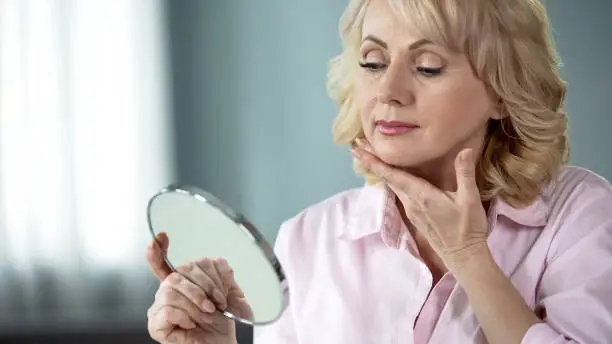 Attractive aged woman looking at her face in mirror, plastic surgery effect