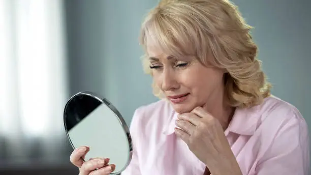 Lady in her 50 looking in mirror remembering young and healthy skin, nostalgia