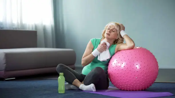 Exhausted active lady wiping with towel after home workout with fitness ball