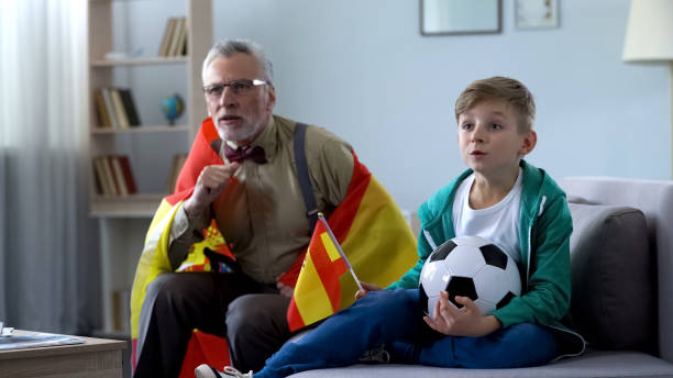 istock Old man wrapped in Spanish flag watching soccer with boy, worrying about game 1072205024
