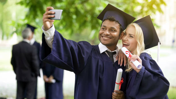 istock Young people in graduation outfit taking selfie with mobile phone, looking at it 1072207316