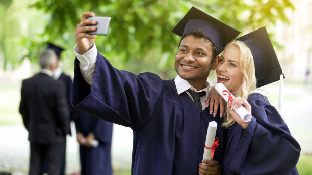 Young people in graduation outfit taking selfie with mobile phone, looking at it Young people in graduation outfit taking selfie with mobile phone, looking at it graduation photos stock pictures, royalty-free photos & images