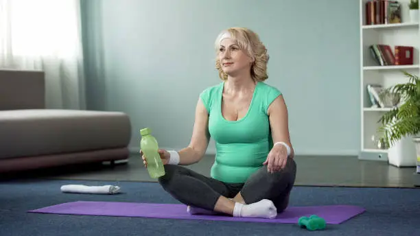 Active aged female sitting on sport mat with bottle of water, workout break