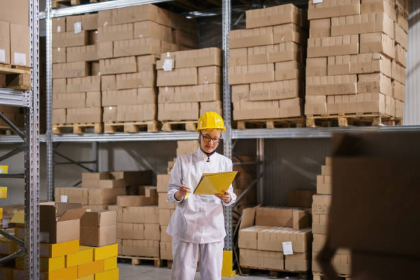 istock Female worker holding folder with paperwork while standing in warehouse. All around boxes on shelves. Protective suit on, helmet on head. 1072244594