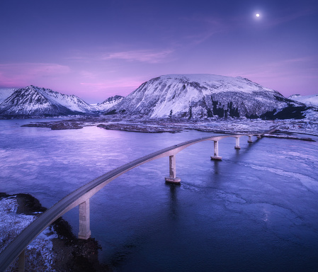 Aerial view of bridge, snow covered mountains, purple sky with clouds and moon, reflection in water. Night landscape with bridge, snowy rocks, blue sea at dusk. Winter in Lofoten islands, Norway. Road
