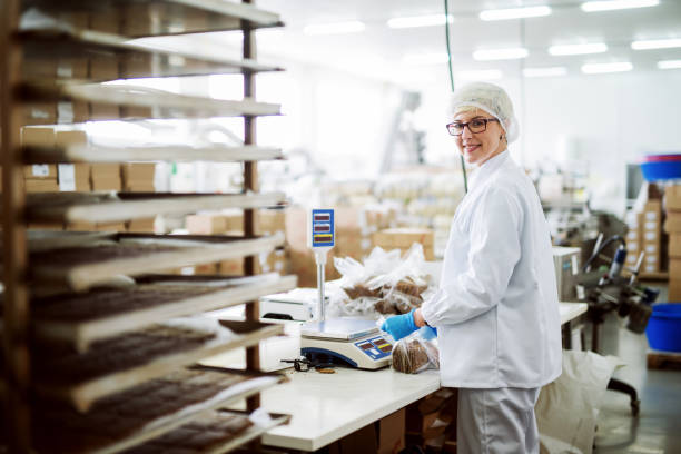 trabajadora midiendo las galletas y ponerlas en bolsas de plástico mientras que de pie y mirando a cámara. interior de la fábrica de alimentos. - quick cookies fotografías e imágenes de stock