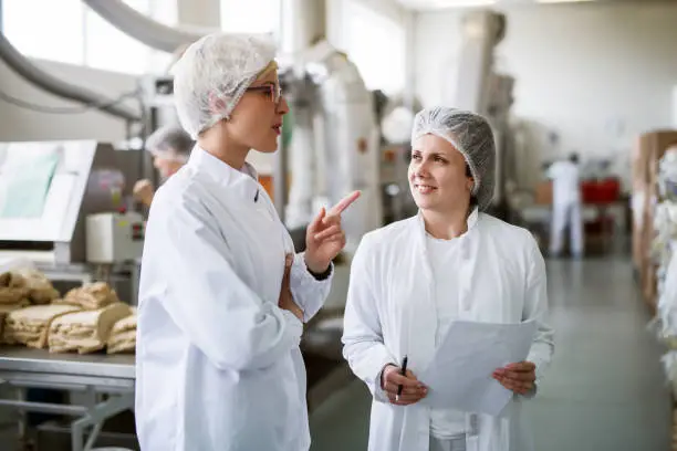 Photo of Two female workers discussing while standing in food factory.