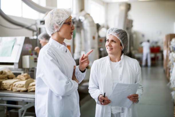 two female workers discussing while standing in food factory. - food safety imagens e fotografias de stock