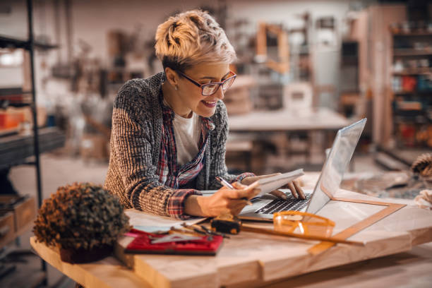 istock Woman using laptop while standing in carpenter workshop. In other hand holding notebook and pen. Female doing male's work concept. 1072252892