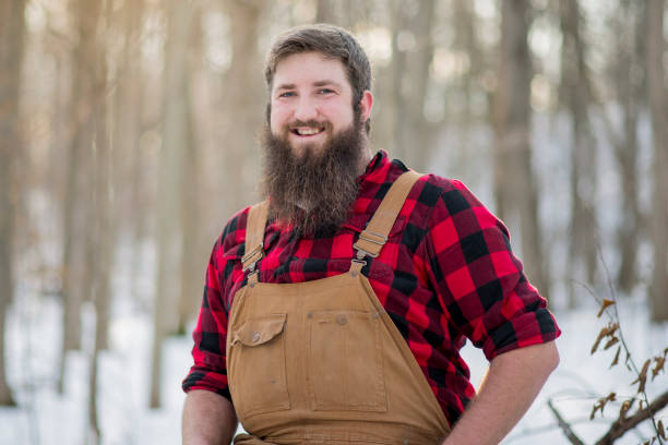 Big Smile An attractive lumberjack man sits in a winter wood. He is bearded, and wears red plaid and brown overalls. The sun shines as the man smiles widely at the camera. man laughing hard stock pictures, royalty-free photos & images