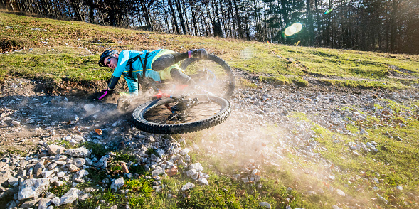 Photo of a mountain biking accident where a woman is falling of her bike on a gravel trail on a meadow in a forest.