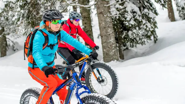 Photo of a woman and a man riding fatbikes on a snowy forest road in the winter.