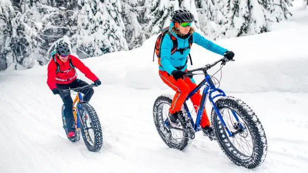 Mountain biker couple riding their fatbikes up a snowy trail in a forest in winter.