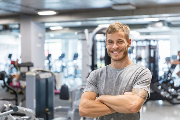 Fitness trainer at gym Portrait of handsome young man standing at gym. Fitness trainer with his arms crossed looking at camera and smiling. fitness trainer stock pictures, royalty-free photos & images