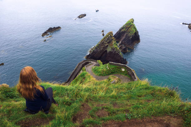 Girl sits on a cliff over the ocean in Ireland Girl sits on a cliff over the ocean and the Dunquin Pier situated on the west coast of the Dingle Peninsula in Ireland dingle bay stock pictures, royalty-free photos & images
