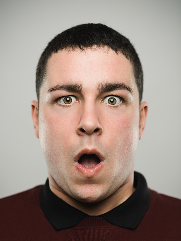 Close-up portrait of shocked real young man with looking at camera. Caucasian male has brown hair and surprised expression. He is against gray background. Vertical studio photography from a DSLR camera. Sharp focus on eyes.