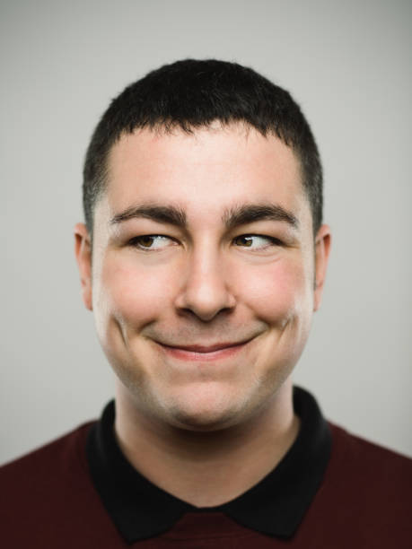 Portrait of a happy young caucasian man looking away. Close-up portrait of real young happy man smirking and looking to the side. Caucasian male has brown hair and naughty expression. He is against white background. Vertical studio photography from a DSLR camera. Sharp focus on eyes. looking around stock pictures, royalty-free photos & images