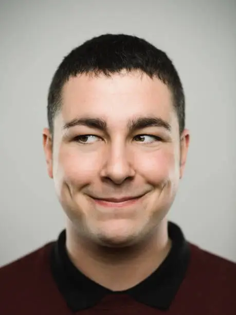 Close-up portrait of real young happy man smirking and looking to the side. Caucasian male has brown hair and naughty expression. He is against white background. Vertical studio photography from a DSLR camera. Sharp focus on eyes.