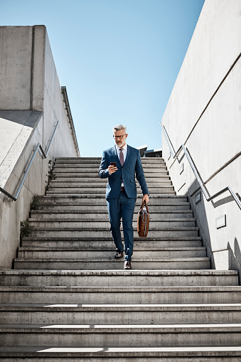 Full length of businessman using mobile phone on steps. Confident professional is moving down staircase against clear sky. Mature executive is wearing suit in city.