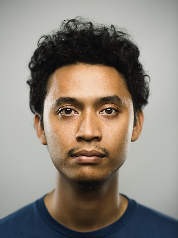 Close up portrait of young adult middle eastern man with blank expression against gray background. Vertical shot of real pakistani man staring in studio with short black hair. Photography from a DSLR camera. Sharp focus on eyes.